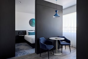 Une chambre d’hôtel moderne, équipée d’un lit, de deux fauteuils, d’une petite table, d’un luminaire rond au plafond et d’une grande fenêtre avec des stores.