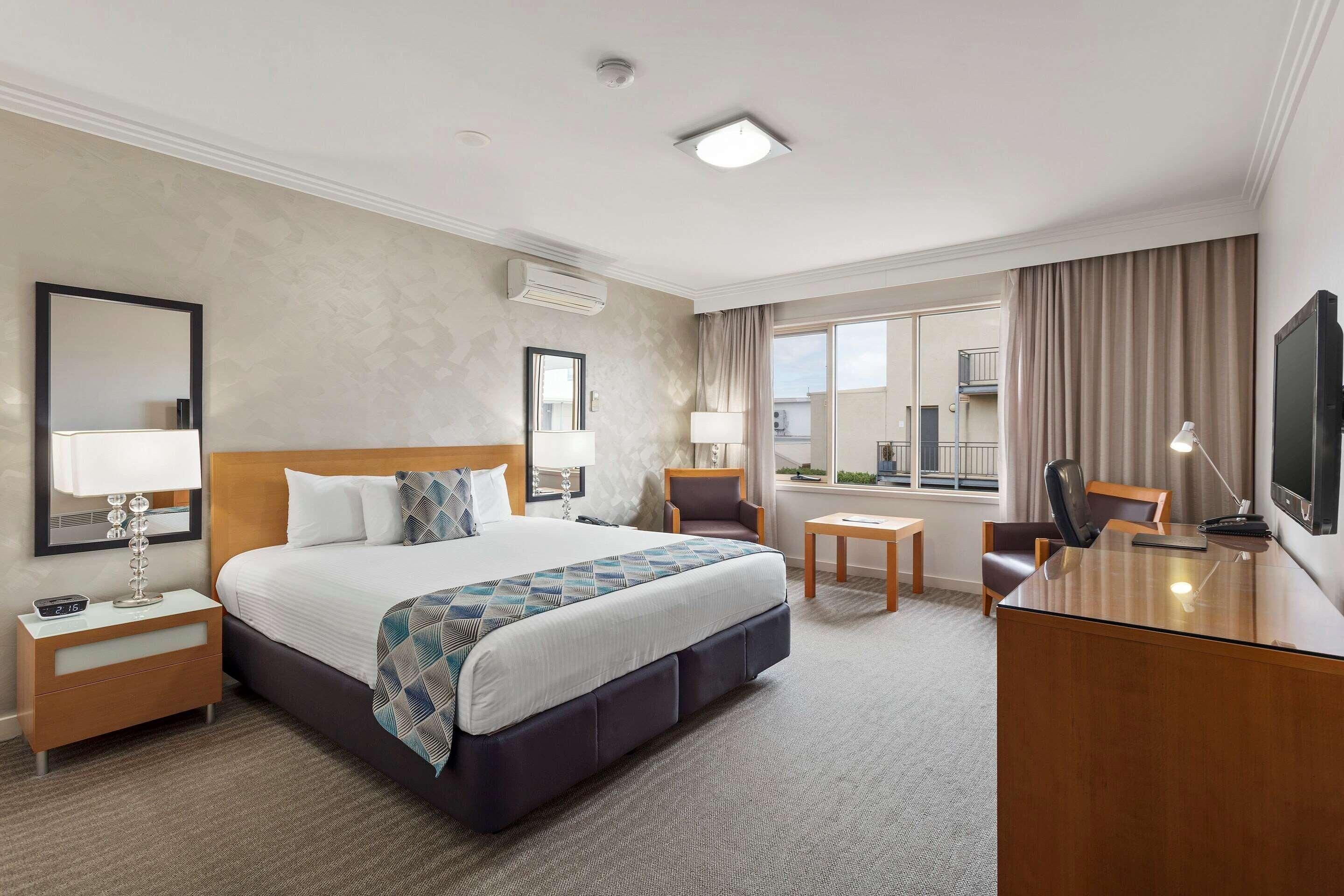 A hotel room with a bed, bedside tables, a desk, a chair, a TV, and a window with curtains.