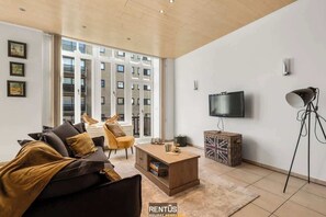 Ein modernes Wohnzimmer mit einer Couch, Couchtisch und Fernseher. Ein großes Fenster bietet Blick auf Gebäude.