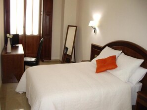 A neatly made bed with white bedding and an orange pillow, a wooden nightstand with a lamp, a wooden desk with a vase of flowers, and a mirror leaning against the wall. - Hotel San Sebastiã¡n Hospederã­a (Cieza)