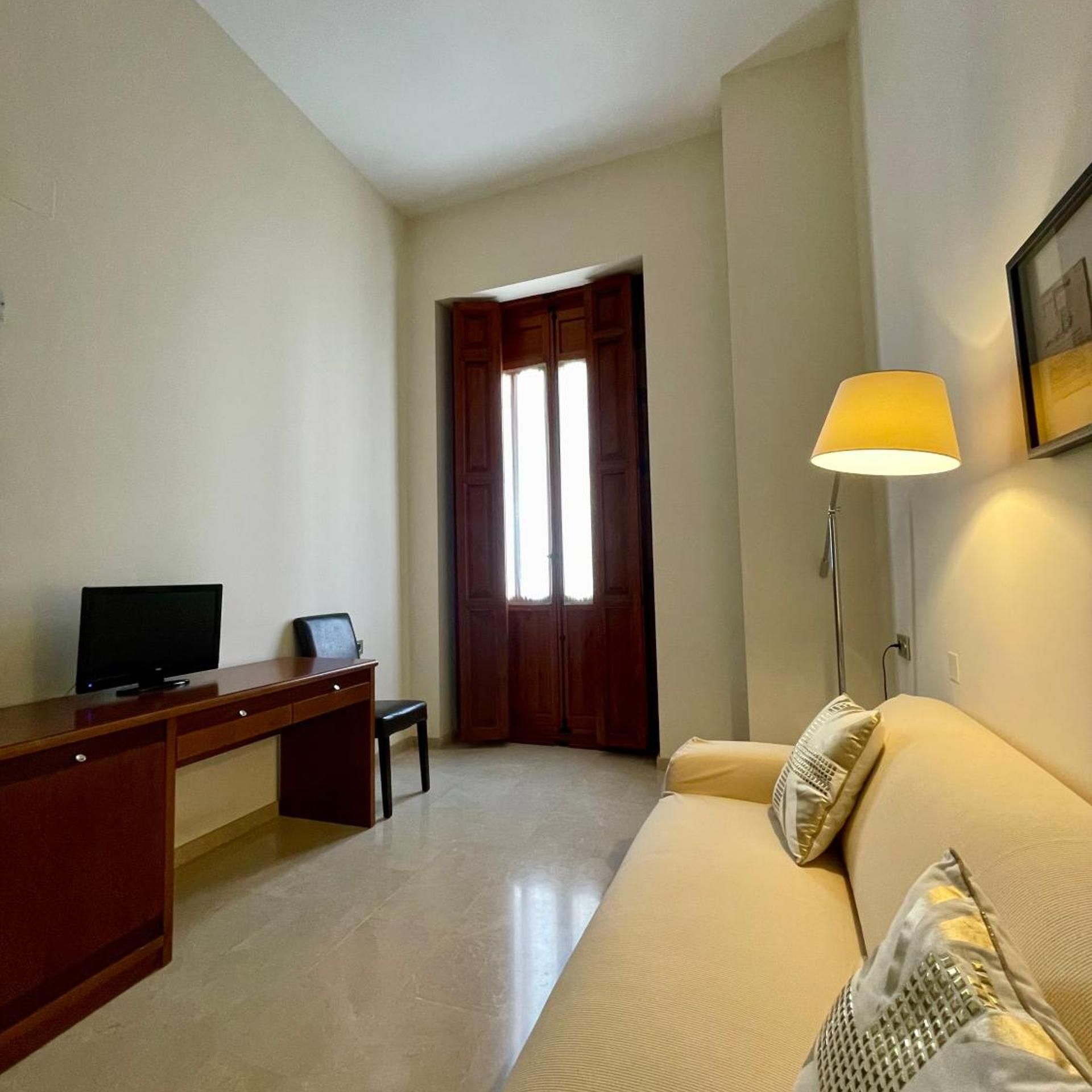 Una habitación con escritorio, silla, televisor, sofá con cojines, lámpara y puerta.