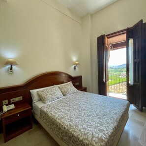 A bedroom with a wooden bed, two bedside lamps, a nightstand with a phone, and a view of a green landscape through an open door. - Hotel San Sebastian Hospederia (Cieza)