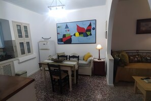 A compact living space with a dining area, a sofa, a small table, a lamp, a refrigerator, and a wall-mounted artwork.