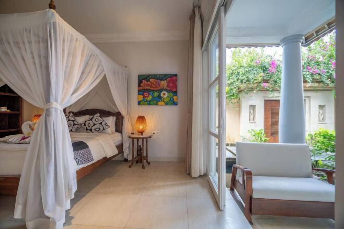 A bedroom with a canopy bed, a bedside table with a lamp, a painting on the wall, and a balcony with a chair.