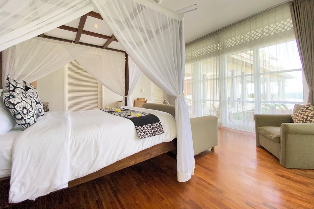 A bedroom with a four-poster bed, a sitting area with a sofa, and a view of the outdoors through large windows.