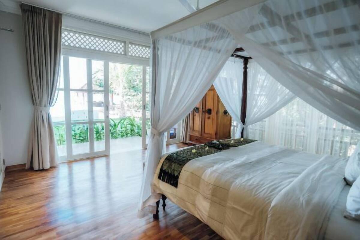 A bedroom with a canopy bed, wooden flooring, and a view of greenery outside.