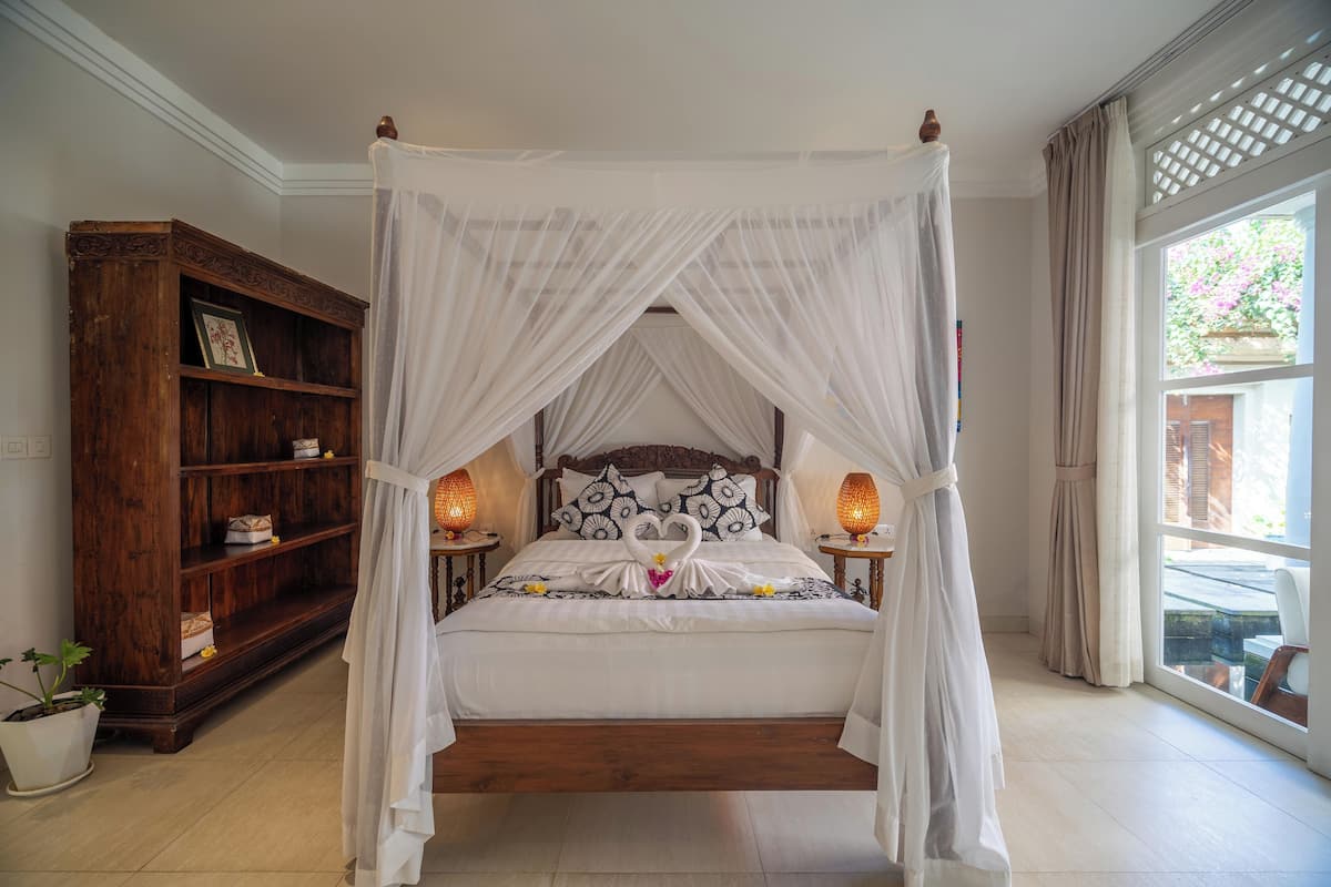 A four-poster canopy bed with a swan-shaped pillow arrangement, a wooden bookshelf, and a large window with a view of greenery.