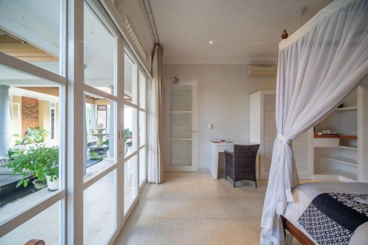 A bedroom with a bed, a desk, and a chair. There is a large window with curtains, and a balcony with plants.