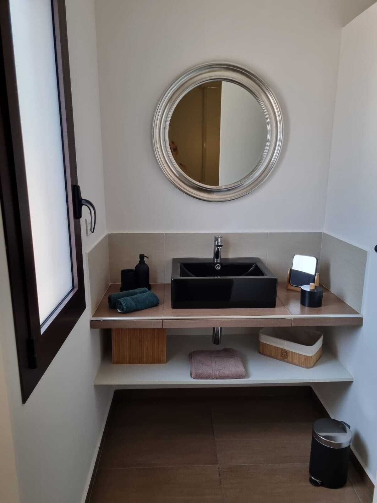 A modern bathroom with a black sink, a round mirror, and a small trash bin.