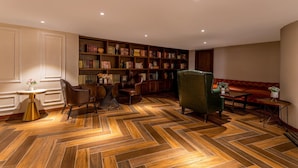 Une pièce dotée d’un parquet en bois à motifs chevrons, d’une bibliothèque, d’un fauteuil vert, d’un canapé en cuir et d’une petite table ronde sur laquelle est posé un vase de fleurs.