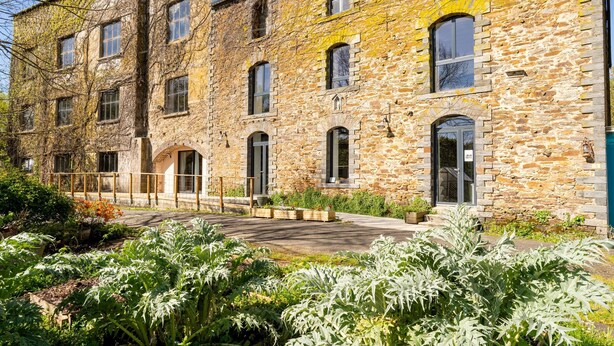 Un edificio de piedra con varias ventanas, un acceso pavimentado y abundante vegetación circundante.