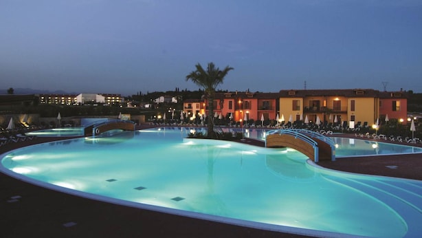 A swimming pool with a bridge, surrounded by buildings with warm lighting, and a palm tree in the center. - Residence Eden Bilo 4 HB (Castelnuovo del Garda)