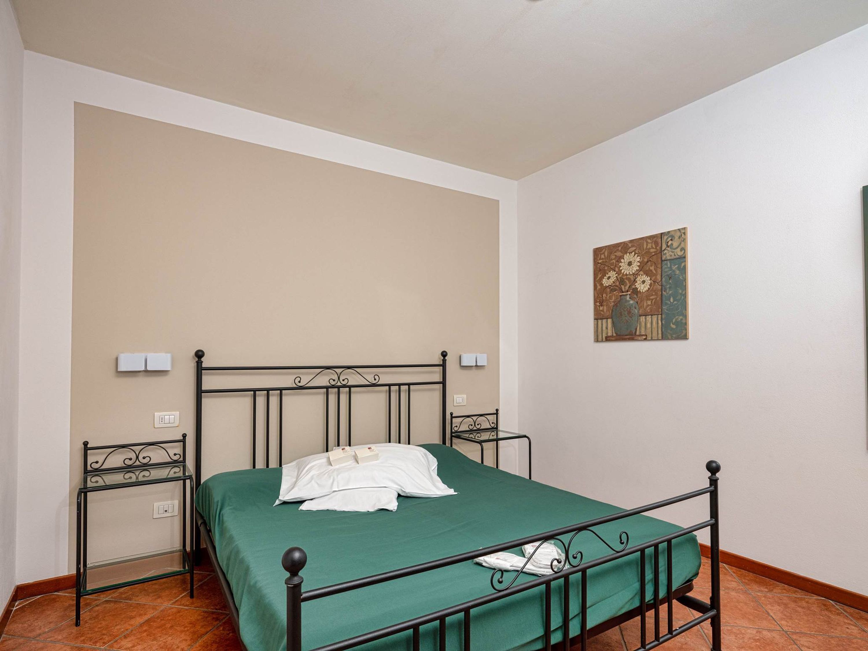 A single bed with a green bedspread, a black metal frame, and a white pillow. There are two bedside tables with white lamps. The room has beige walls and a tiled floor.