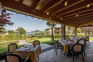 An outdoor dining area with tables and chairs, a stone building in the background, and a view of the sea. - Residence Onda Blu Bilo BB (Manerba del Garda)