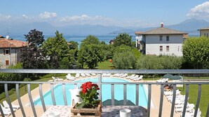 A pool with lounge chairs, a table with a flower, and a view of the sea and mountains. - Residence Nettuno Bilo (Peschiera del Garda)