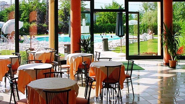 An indoor dining area with tables and chairs, large windows, and a view of an outdoor pool area. - Residence Nettuno Bilo (Peschiera del Garda)