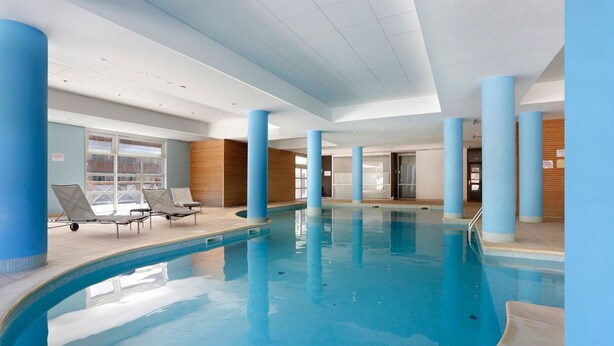 An indoor swimming pool with blue columns, lounge chairs, and a view of the outside through large windows. - Les Chalets du Verdon 2p5n (Allos)