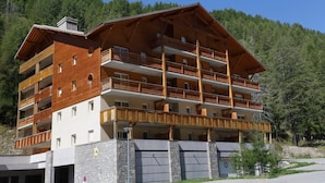 A multi-story hotel with balconies, a stone base, and a forested backdrop. - Les Chalets du Verdon 2p5n (Allos)