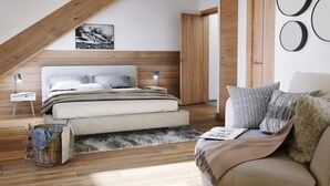 Un dormitorio con techo inclinado, una cama con cabecera, una mesita de noche, una lámpara, una cesta de mimbre, un sofá y una puerta.
