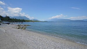 En strand med småsten och en brygga som sträcker sig ut i stilla vatten, omgiven av berg och klarblå himmel.