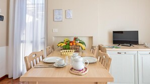 A dining area with a wooden table set for two, a fruit basket, and a television in the background. - Residence Eden Bilo 4 BB (Castelnuovo del Garda)