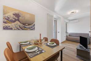 A dining area with a wooden table set for four, a painting of a wave, and a modern living space with a sofa and TV. - Residenza I 3 Mori B (Arco)