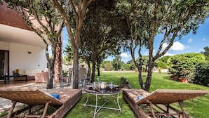 Eine Terrasse mit Korlounge-Stühlen, einem Tisch mit Topf und Gläsern und Blick auf einen üppigen Garten und ein Gebäude.