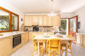 A kitchen with wooden cabinets, a dining table with wicker chairs, and a view of greenery outside. - Casa Ortensia 2 in Arzachena (Arzachena)