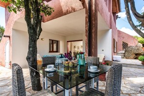 An outdoor dining area with a glass table and wicker chairs, surrounded by stone flooring and a large tree. - Casa Ortensia 2 in Arzachena (Arzachena)