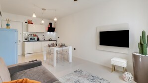 A modern living room with a grey sofa, a coffee table, and a dining area with a white table and chairs. A ceiling fan and pendant lights are visible.