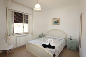 Ein Schlafzimmer mit Bett, Stuhl, Nachttisch, Lampe, Heizkörper, einem Fenster mit Jalousien und einem Bild an der Wand.