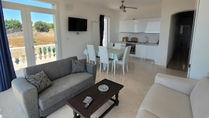 A modern living room with a sofa, a coffee table, and a TV. There is a dining area with a table and chairs, and a kitchen with white cabinets and a ceiling fan. - Felloniche Apartment Sea View (Castrignano del Capo)