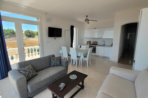Ein modernes Wohnzimmer mit einer Couch, einem Couchtisch und einem Fernseher. Es gibt eine Essecke mit Tisch und Stühlen sowie eine Küche mit weißen Schränken und einem Deckenventilator.