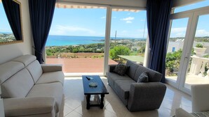 A living room with a beige sofa, a grey sofa, and a coffee table. There is a large window with a view of the sea and greenery. - Felloniche Apartment Sea View (Castrignano del Capo)