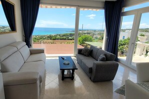 Ein Wohnzimmer mit einem beigen Sofa, einem grauen Sofa und einem Couchtisch. Ein großes Fenster bietet Blick auf das Meer und viel Grün.