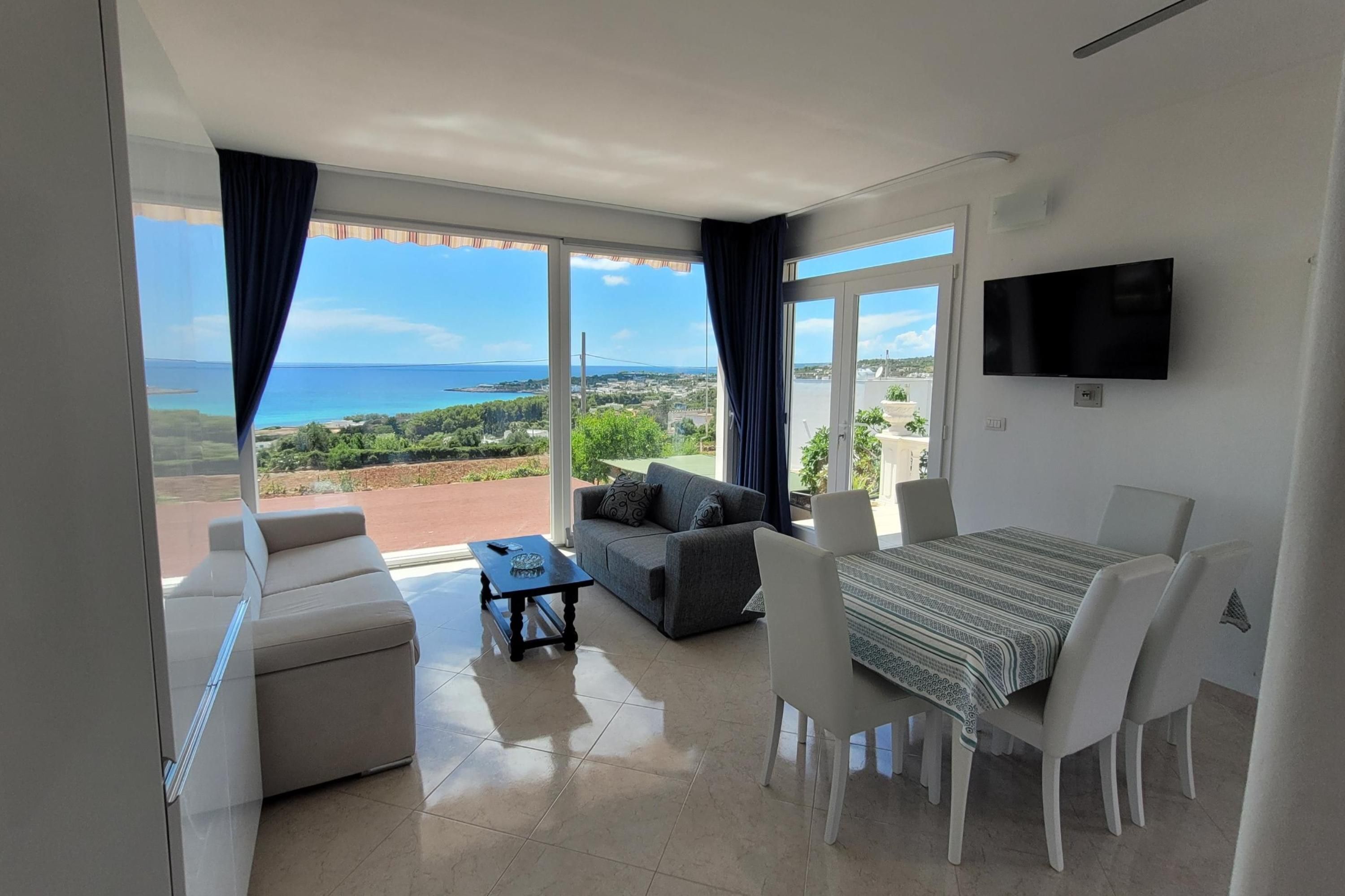 Una sala de estar moderna con un sofá, una mesa de comedor y un televisor. Una ventana grande ofrece vistas al mar y al paisaje.