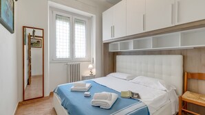 Siististi pedattu sänky, jossa on valkoiset lakanat ja sininen peitto, puinen tuoli ja pieni yöpöytä, jossa on lamppu.