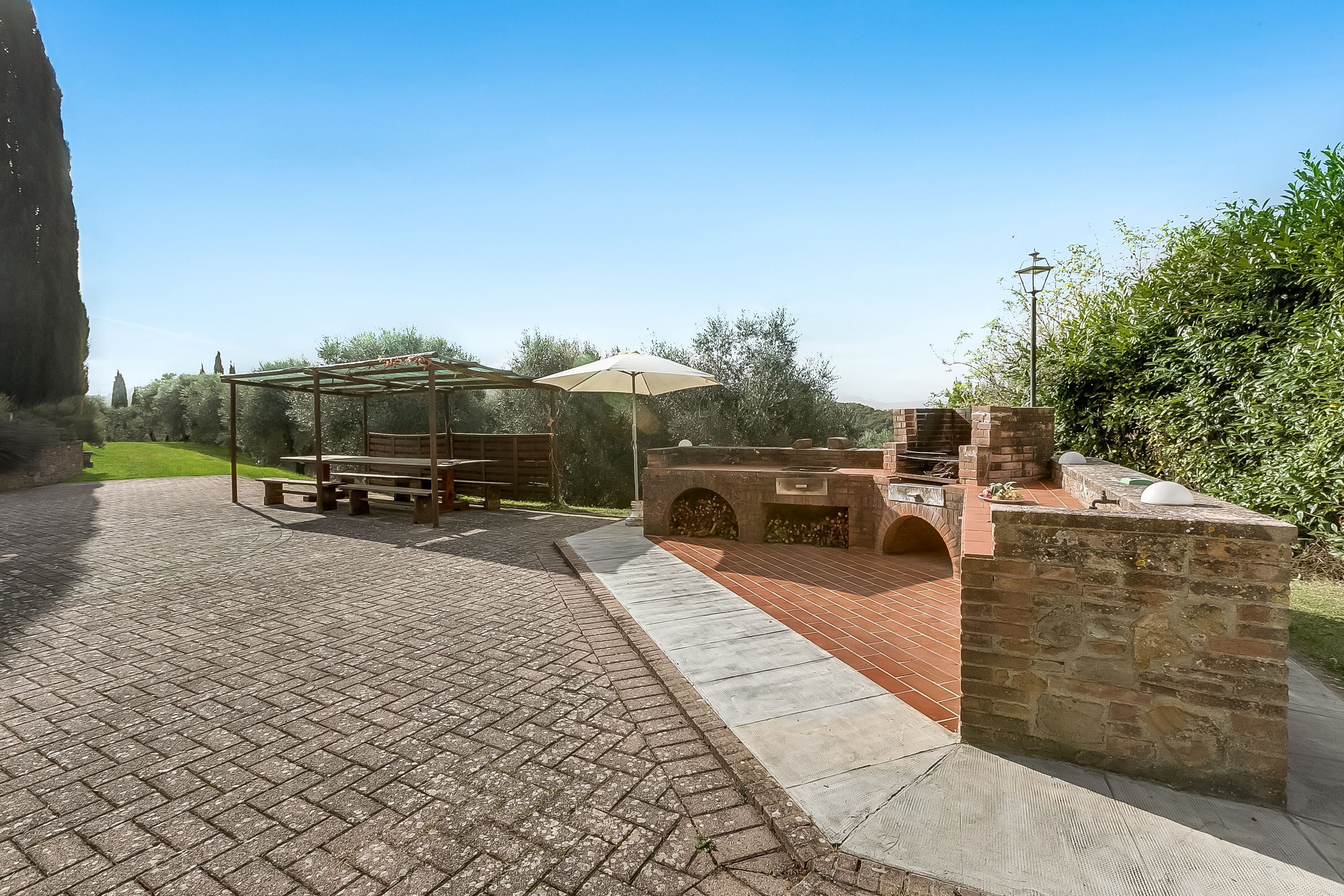 An outdoor patio with a brick floor, a wooden bench, a pergola with an umbrella, and a stone barbecue area.