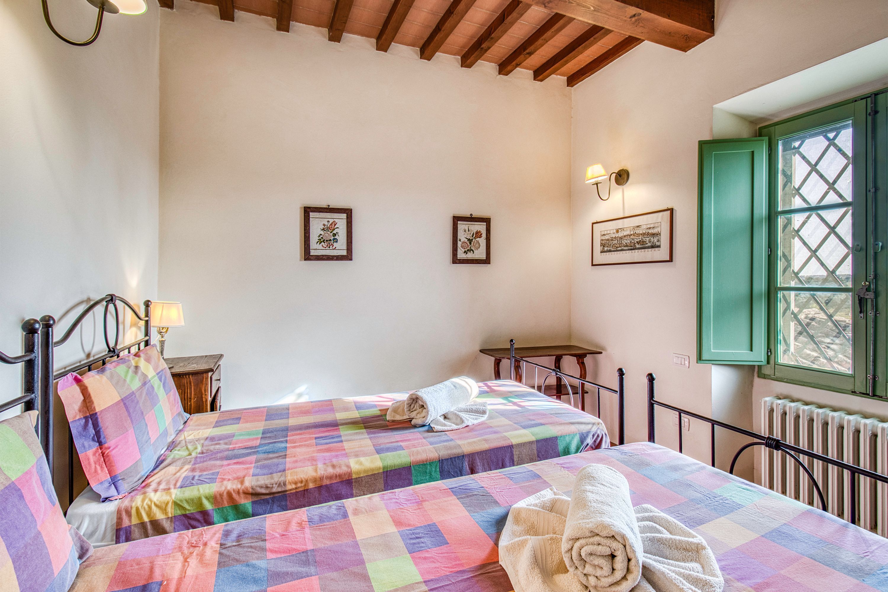 Un dormitorio con dos camas, una ventana con contraventanas verdes y cuadros enmarcados en la pared.