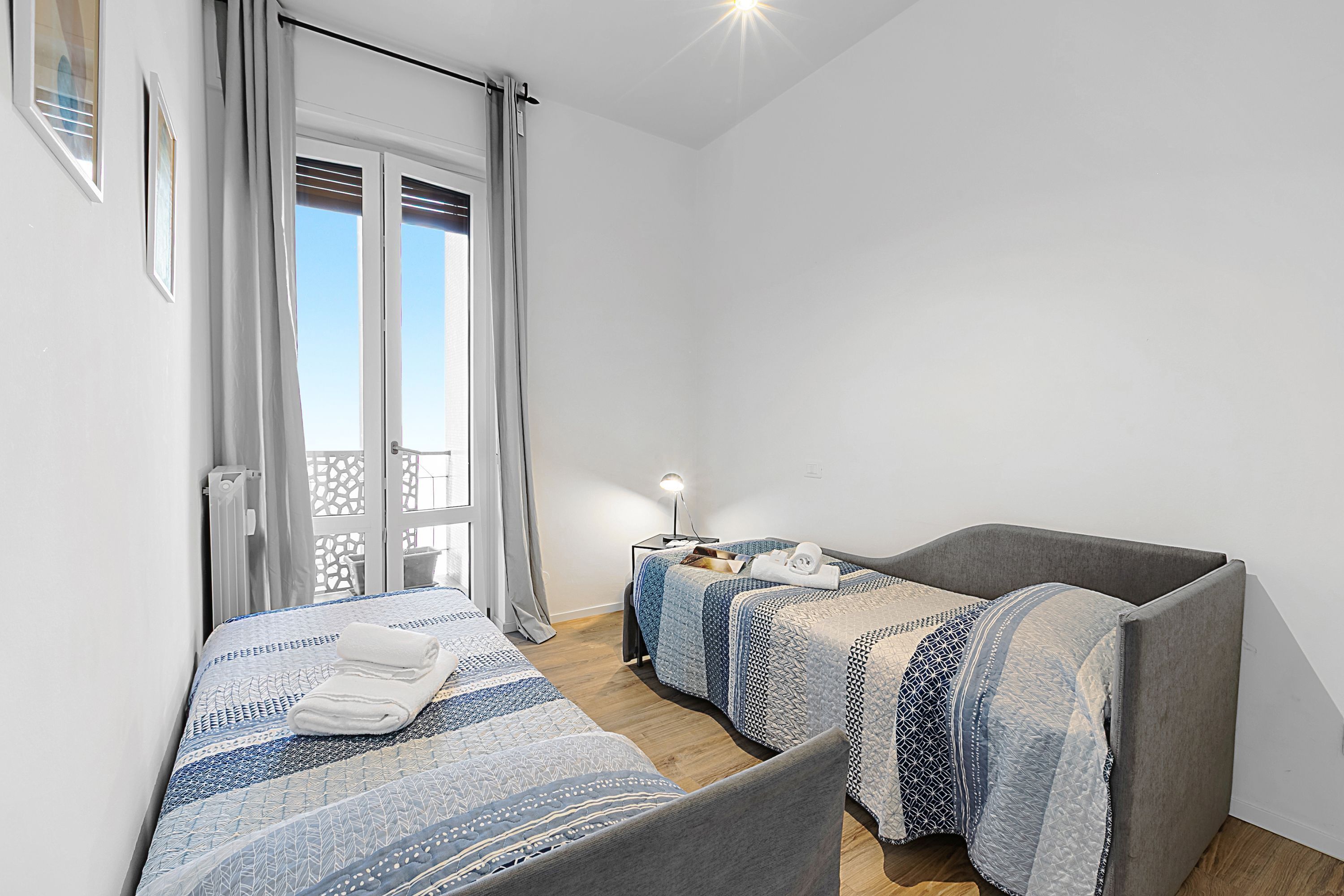 A room with two beds, a small table with a lamp, and a balcony with curtains.