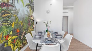 A dining area with a tropical mural, white chairs, a round table set with plates, glasses, and a vase with flowers. - Specchio Di Cielo Arona Center (Arona)