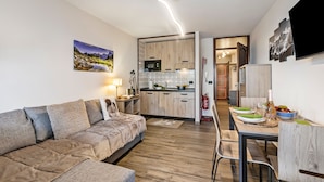 A modern living room with a sofa, a dining table with chairs, a kitchen area with wooden cabinets, and a mounted flat-screen TV. - Prestige Eleven In The Center Of Town (Sestriere)