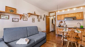 Um espaço de estar compacto com sofá cinza, mesa de jantar de madeira e cadeiras, cozinha com armários de madeira e painel de azulejos, e prateleiras com molduras e quadros.