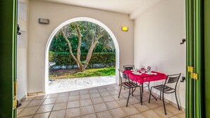 A room with a tiled floor, a table with a red tablecloth, two chairs, and an arched doorway leading to a garden with trees and a fence. - Family Village M7 Pool And Sea (Otranto)