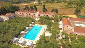 A large swimming pool surrounded by lounge chairs and umbrellas, with a backdrop of a large building complex and surrounding greenery. - Family Village M7 Pool And Sea (Otranto)