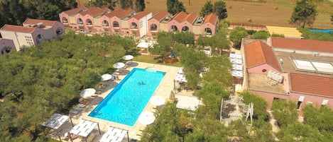 A large swimming pool surrounded by lounge chairs and umbrellas, with a backdrop of a large building complex and surrounding greenery.