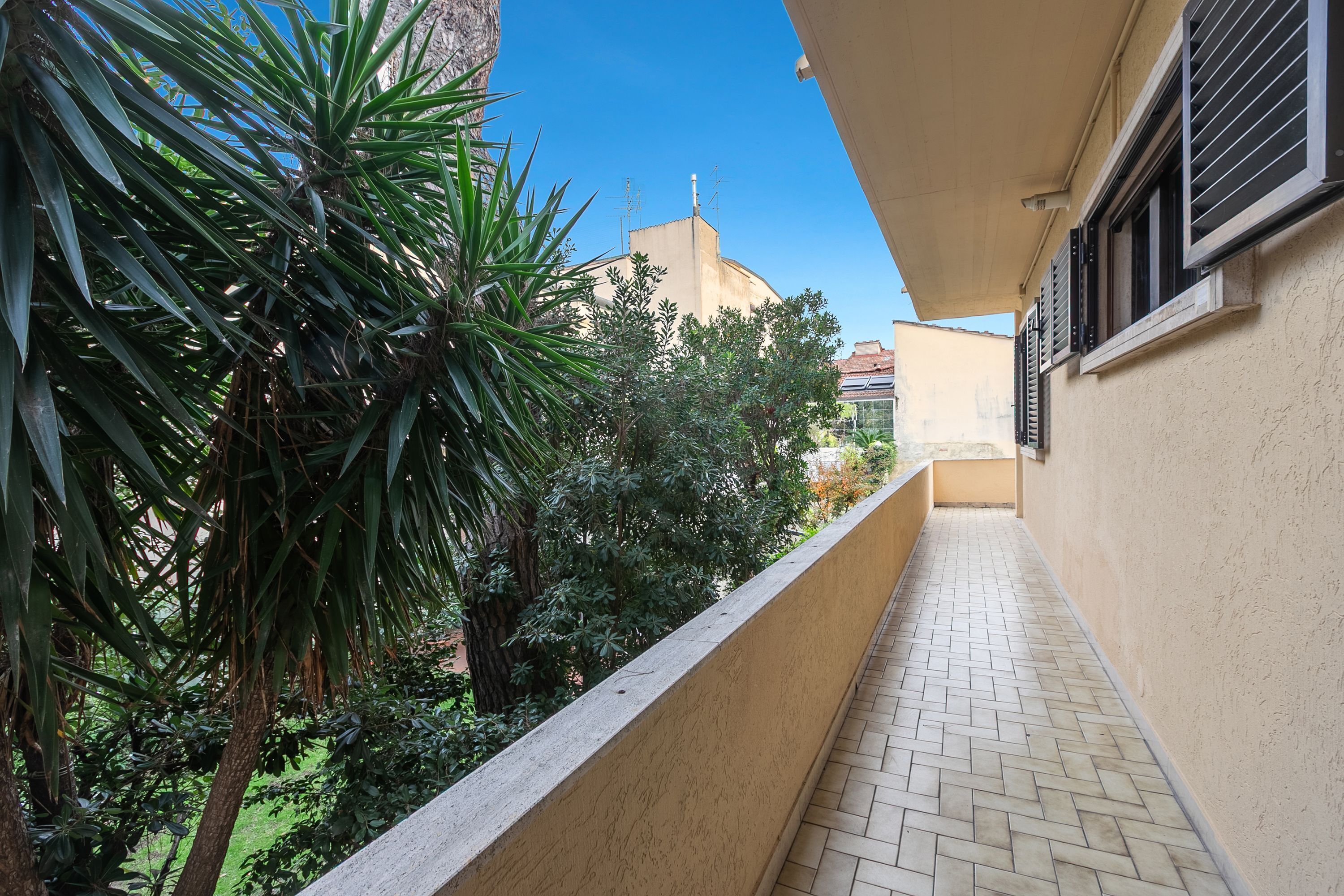 A narrow corridor with a tiled floor, surrounded by greenery and a building with a beige facade.