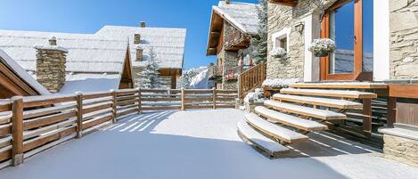 Un espace extérieur enneigé, avec des bancs en bois et un bâtiment en pierre doté d’un balcon en bois.