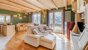 A spacious room with wooden floors, a white sofa, a dining table with chairs, and a view of mountains through large windows. - Baita Delle Stelle in Scarlino (Sauze di Cesana)