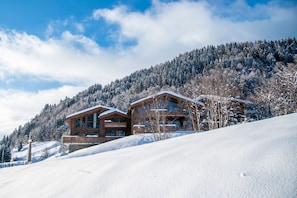 A wooden chalet with multiple balconies, situated on a snow-covered slope with a forested mountain backdrop. - R Sidence Les Portes de Meg ve C58e (Praz-sur-Arly)
